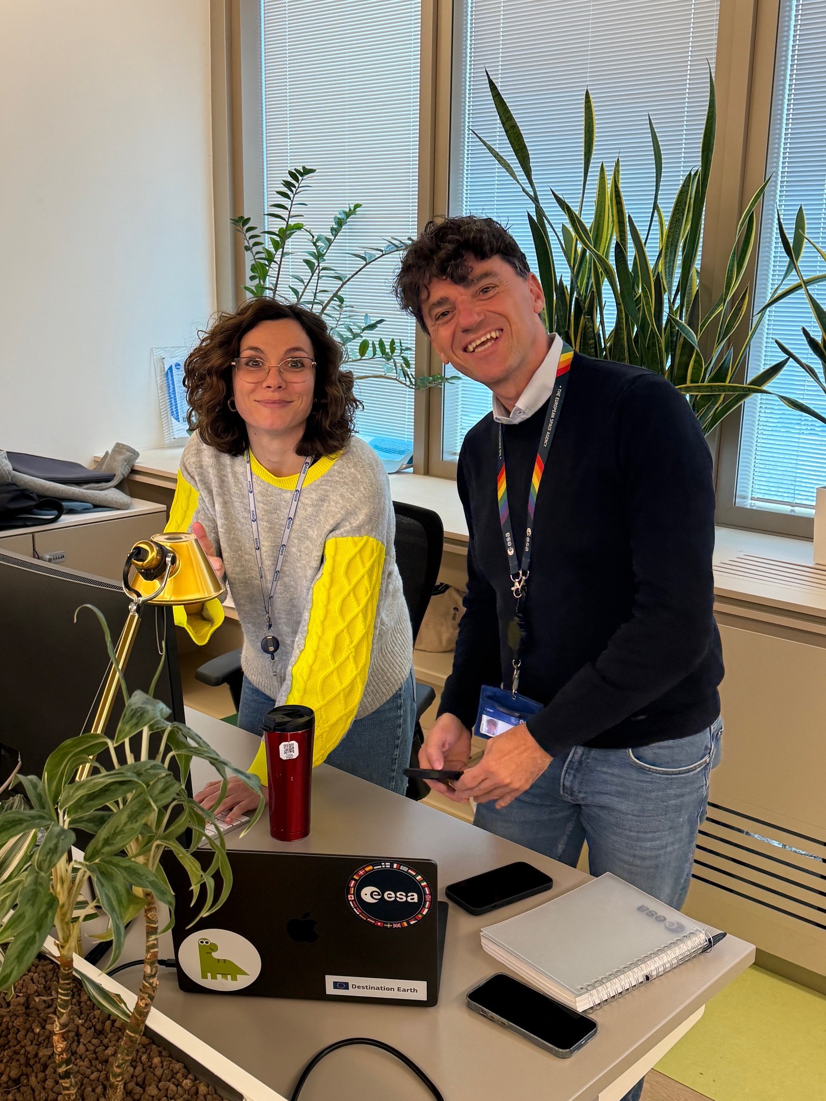 A man (Luca Girardo) and a woman (Barbara Borgia) are in an office. They face the camera and smile. They are both standing near a desk.